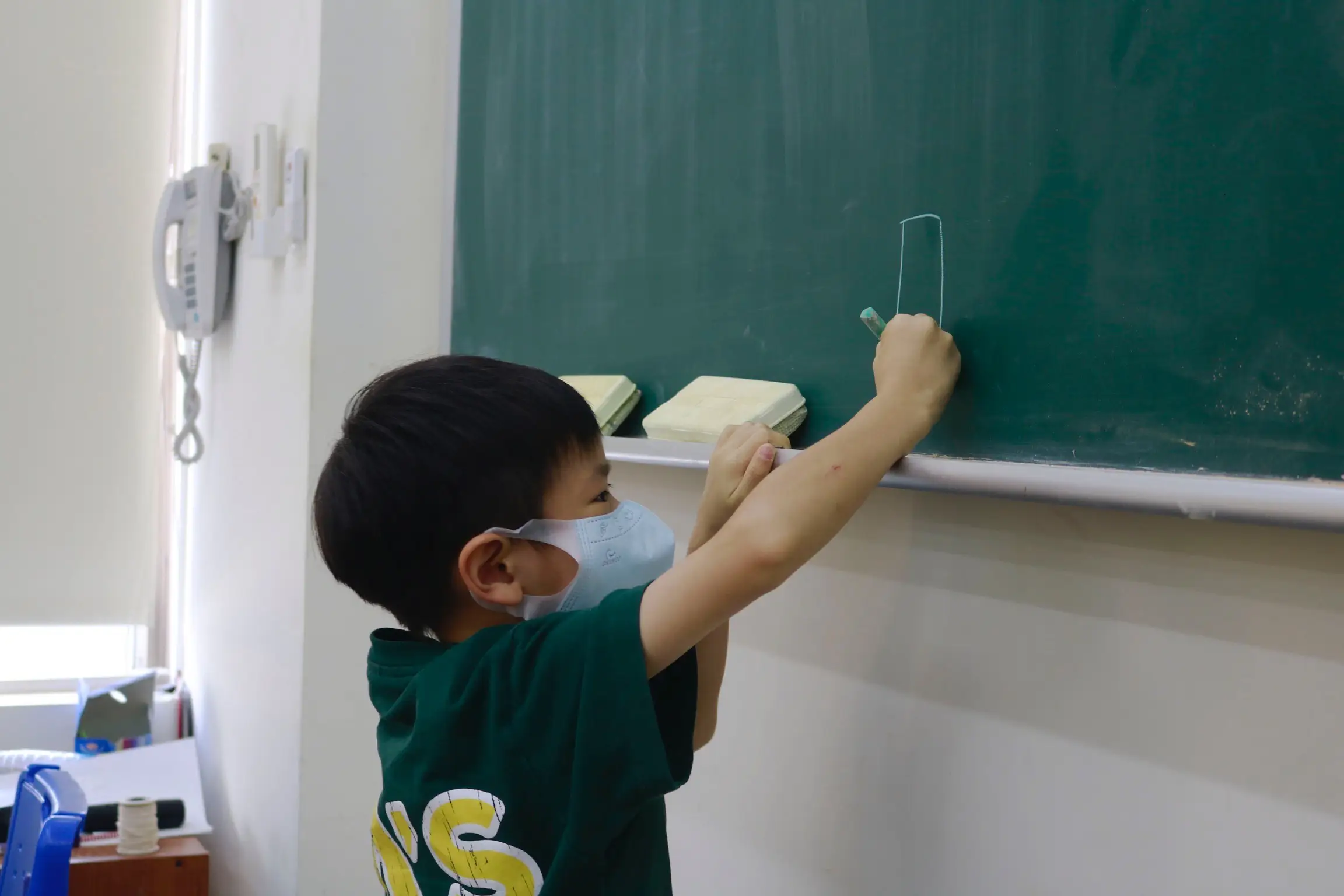 一個戴口罩的孩子正在黑板上書寫的特寫照片。
