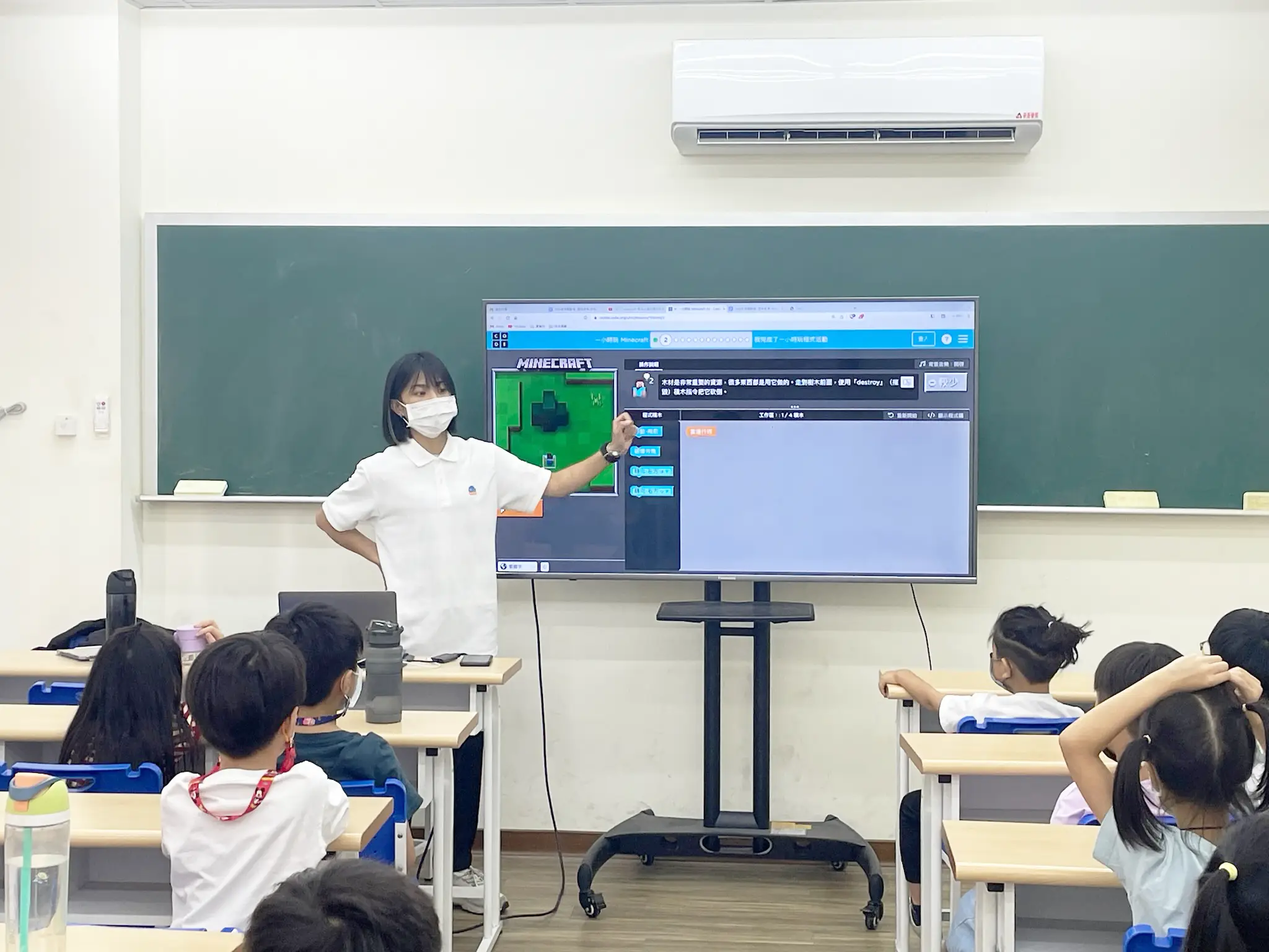 一位老師正在指著投影布幕上的Minecraft操作介面進行講解。